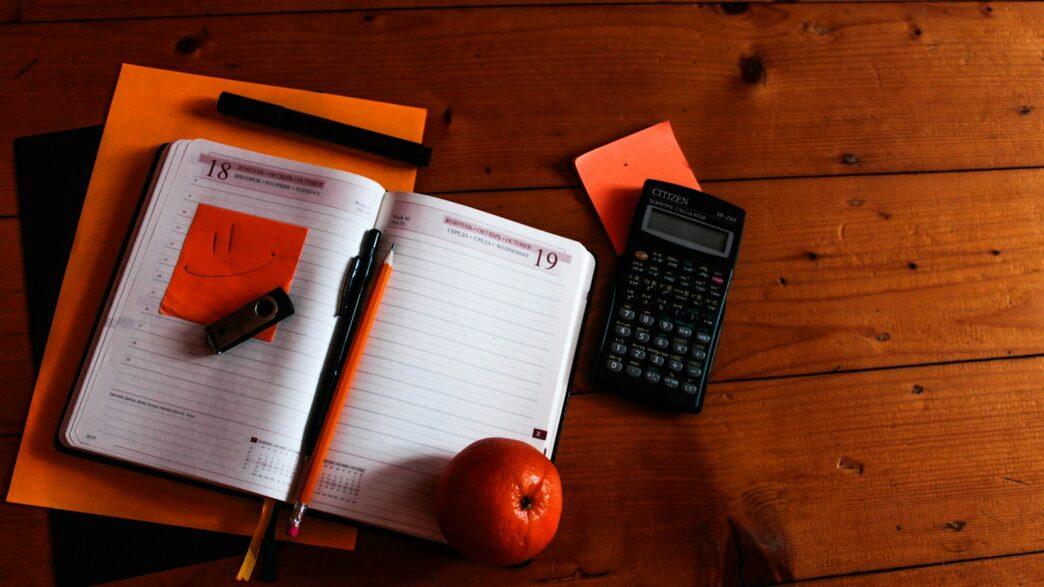 black scientific calculator, orange, and notebook on table