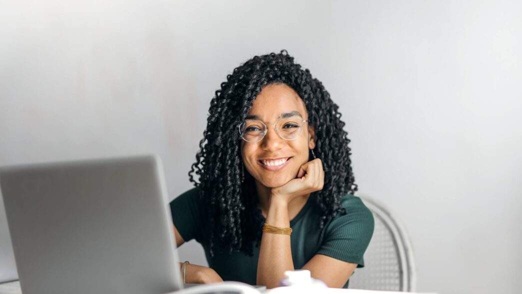 Joyful businesswoman with curly hair smiling at camera while using laptop indoors.